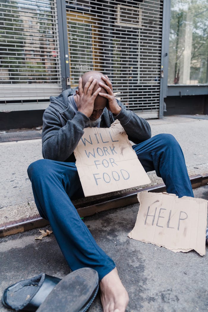 A man sits on a city sidewalk holding a sign that reads 'Will Work for Food' reflecting poverty and unemployment.